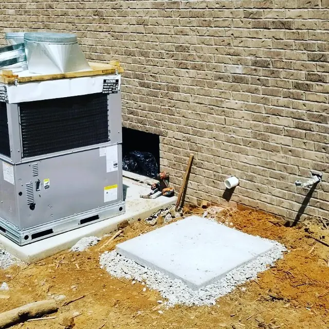 New package HVAC unit set on a concrete pad beside a new brick home, ductwork fittings staged on top